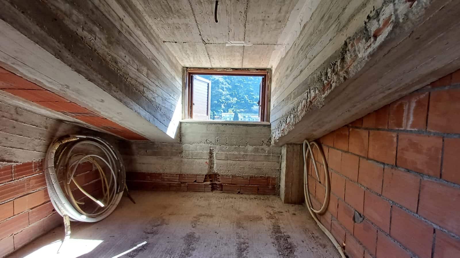 Finestra in mansarda in costruzione con vista sulle montagne, ambiente da completare, struttura in cemento e mattoni rossi, ideale per progetti di ristrutturazione e case personalizzate.