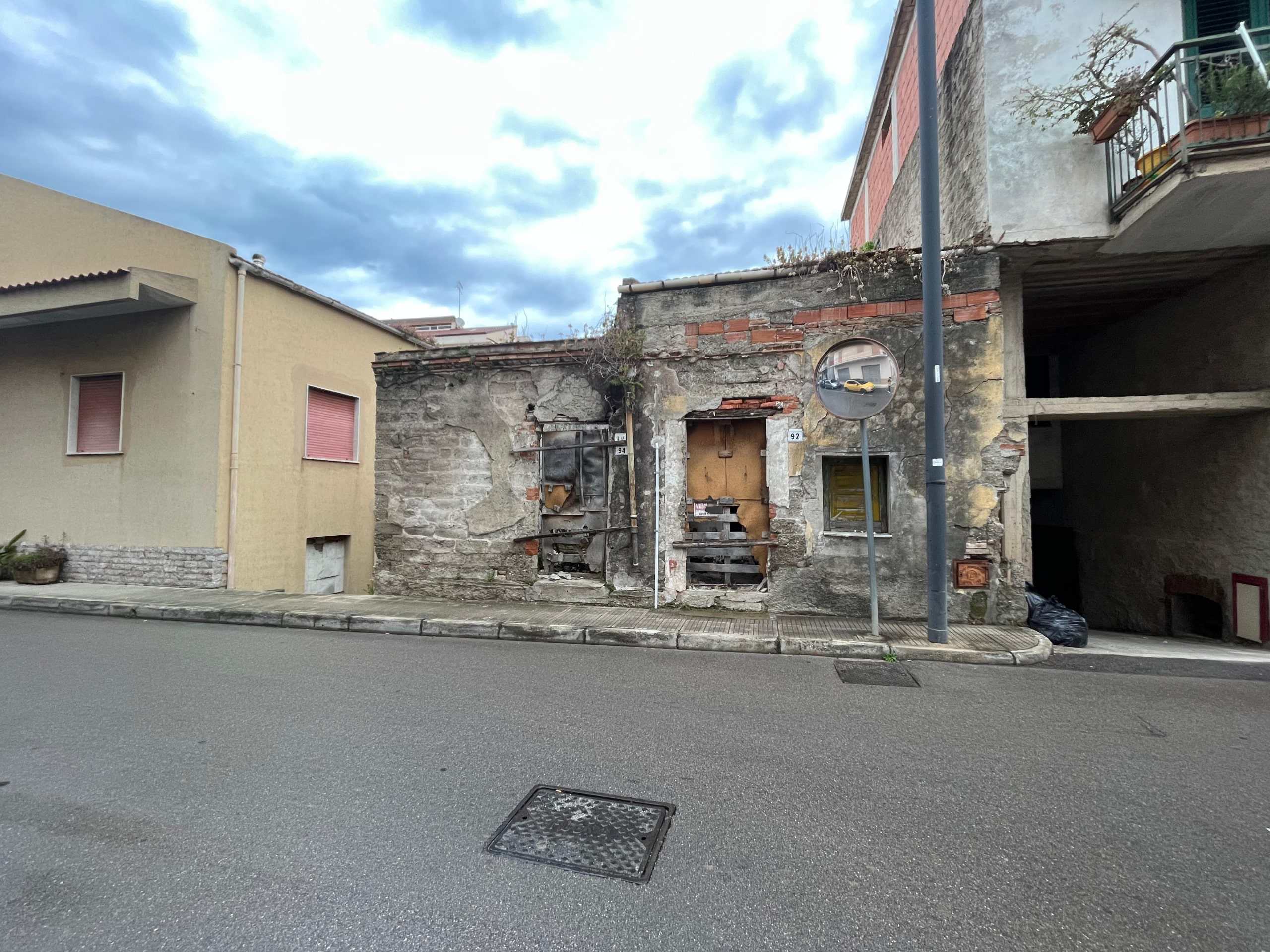 Vecchio edificio in rovina con pareti in pietra e mattoni, situato su una strada urbana, con alcuni elementi di abbandono e deterioramento, accanto a strutture più recenti e moderne.
