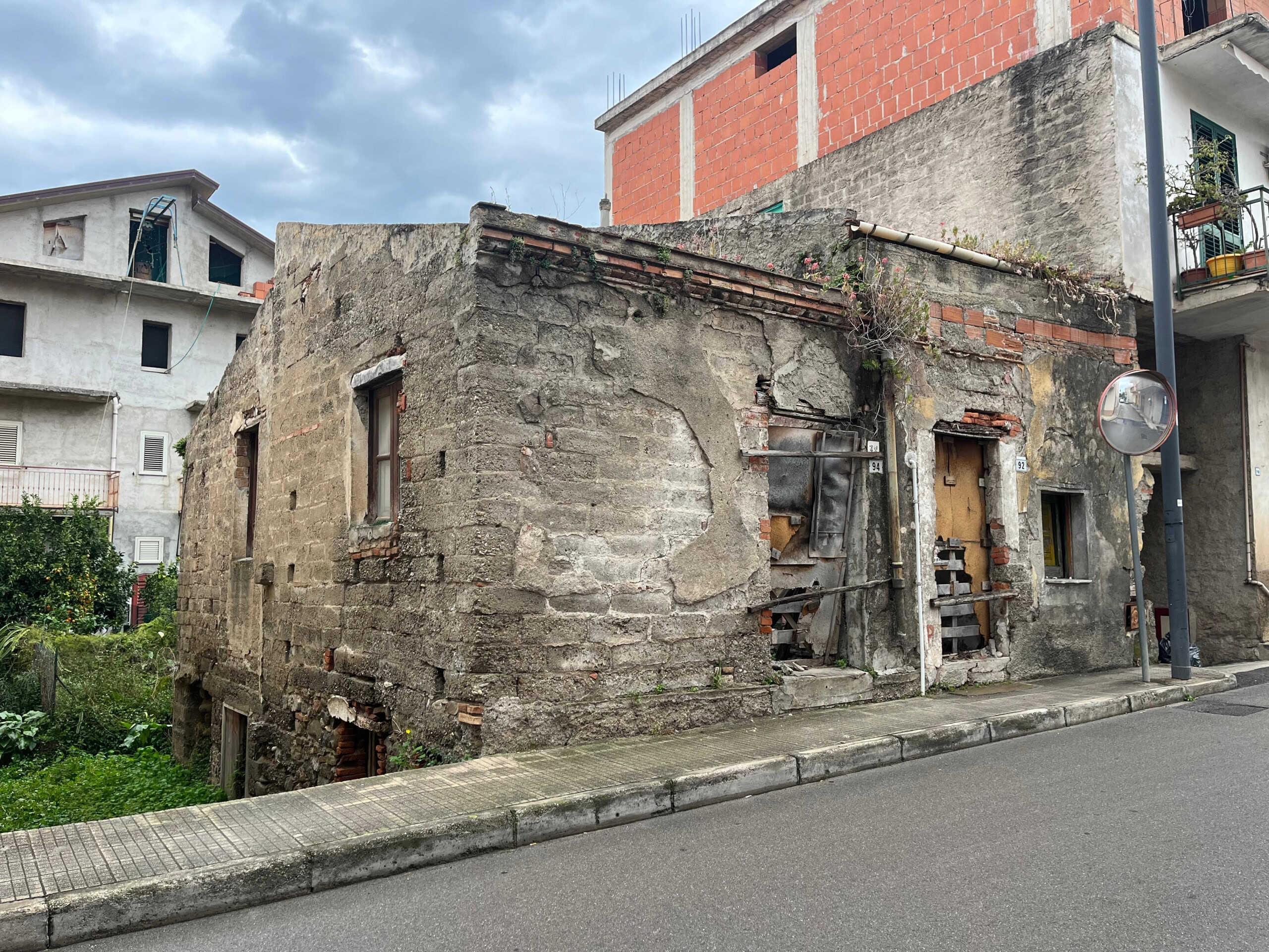 Vecchia casa abbandonata in zona urbana con muri di pietra e pareti in mattoni rossi, portoni e finestre in condizioni di deterioramento, scenario di edilizia storica in stato di abbandono.