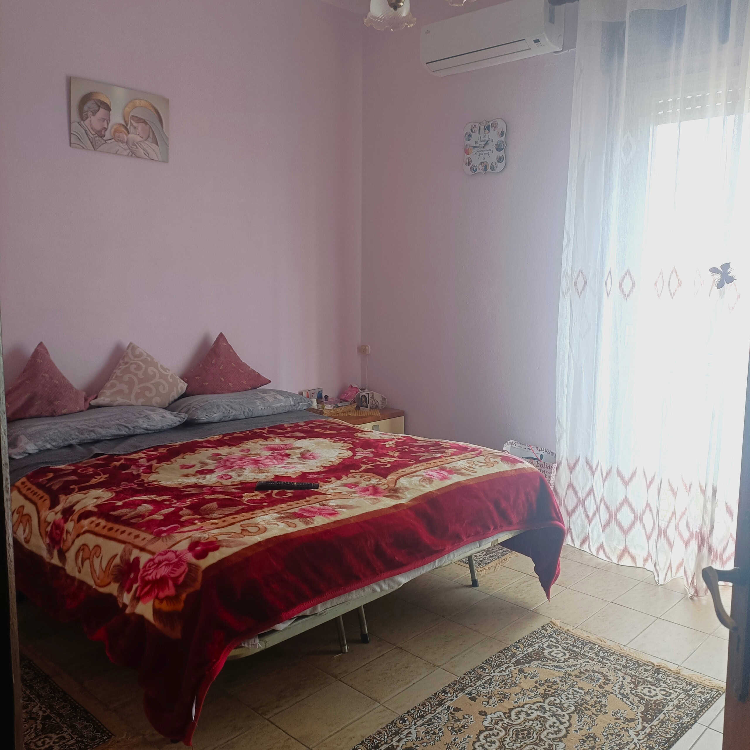 Letto matrimoniale in camera da letto con lenzuola colorate, decorazioni e quadri sacri, ambiente luminoso con aria condizionata e tende trasparenti.
