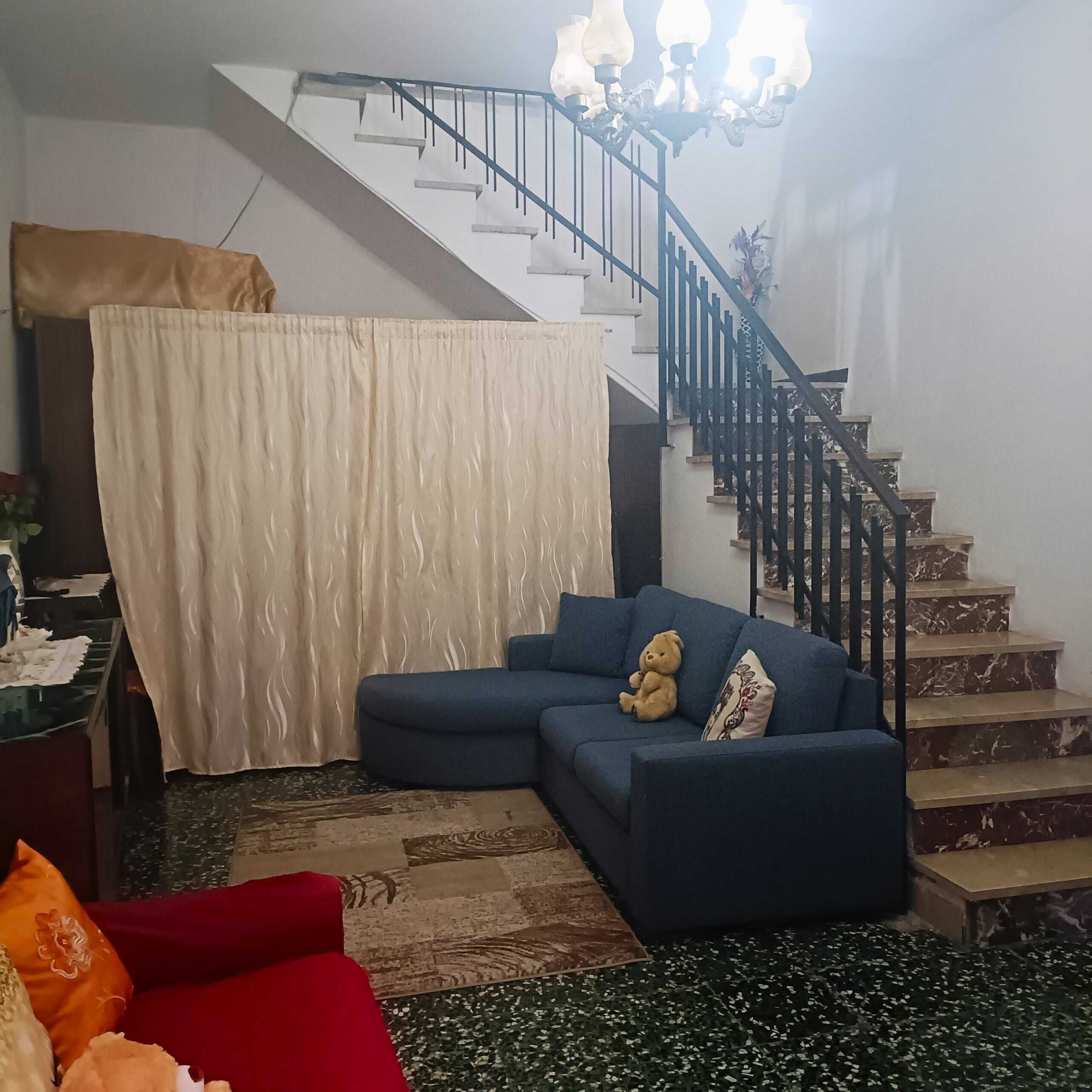 Divano blu in soggiorno con teddy bear e cuscino decorativo, scala in marmo con ringhiera nera, lampadario a braccio in stile classico, arredamento accogliente, NextCasa interior design.