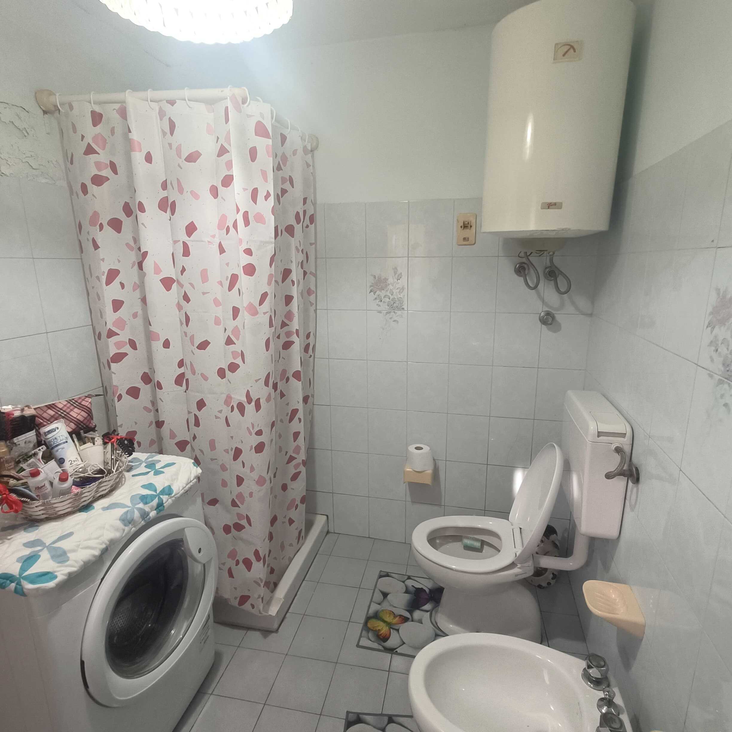 Lavanderia bagno con lavatrice, lavabo, WC, bidet, bollitore e doccia con tende floreali, ambiente funzionale in stile semplice e pratico, ideale per piccoli appartamenti o case.