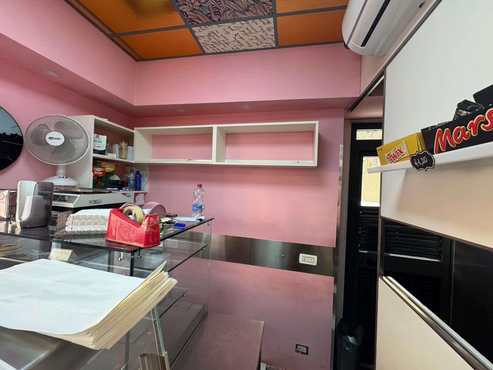 Finestra di un negozio o ufficio con pareti rosa, scaffali aperti, un ventilatore da tavolo e vari prodotti alimentari sugli scaffali, ambiente commerciale accogliente e colorato.