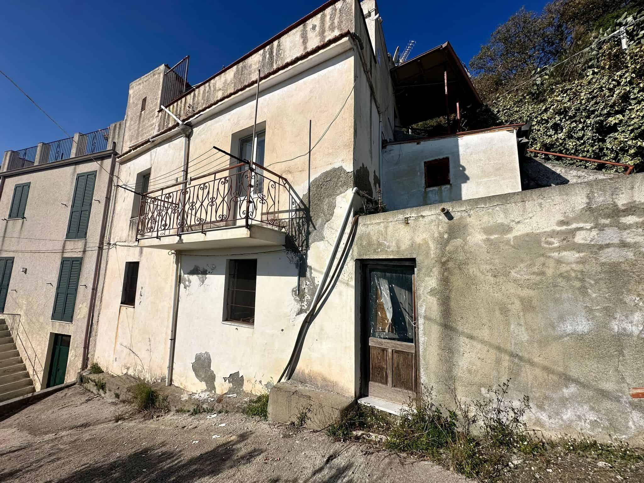 Vecchio edificio residenziale con pareti danneggiate e balcone in ferro battuto, situato su una strada in pendenza con chiostro e natura circostante, caratteristiche tipiche di immobili da ristrutturare.
