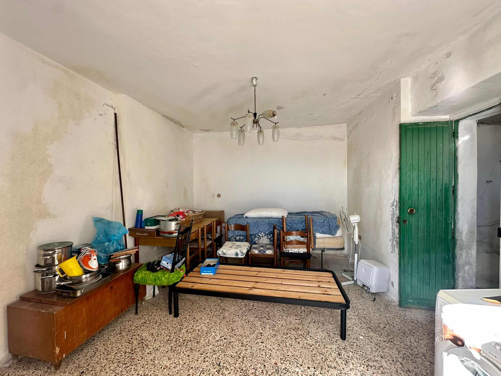 Vecchio soggiorno con pareti in cattivo stato e arredamento semplice, in attesa di ristrutturazione, con asciugacapelli, tavolo in legno e sedie, e porta verde a pannello.