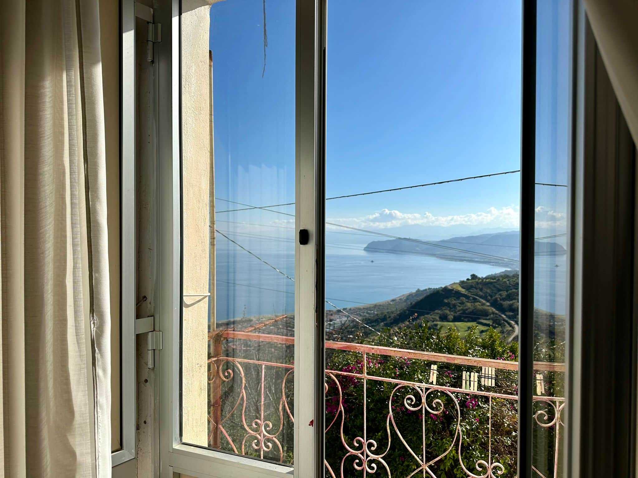 Vista panoramica dal soggiorno con finestra aperta, mostrando il mare, le colline e un cielo sereno, ideale per promuovere appartamenti con vista mare e location esclusive.
