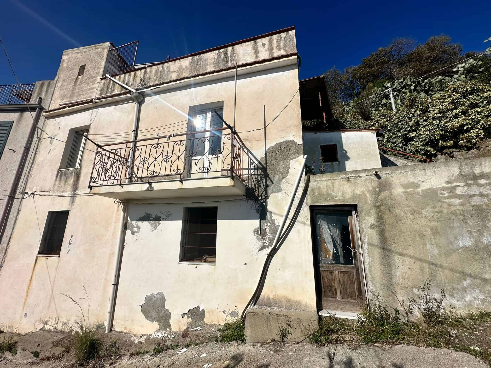 Vecchia casa in condizioni di abbandono, struttura in pietra e cemento con balcone in ferro battuto, pareti rovinate e infissi in legno, situata in zona collinare soleggiata con vegetazione sullo sfondo.
