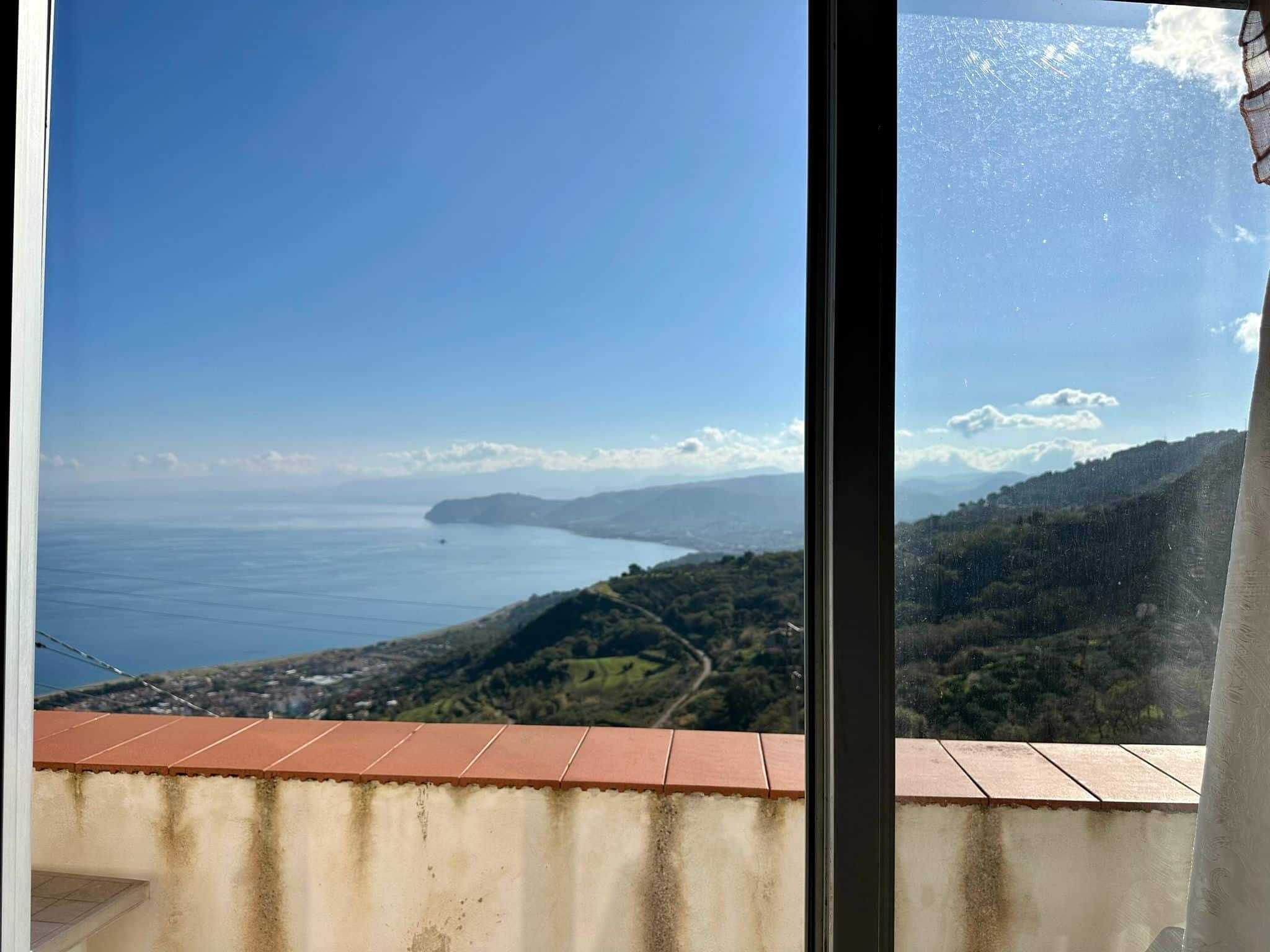 Vista panoramica mare e colline dal soggiorno, grande finestra con vista mozzafiato sulla costa e paesaggi naturali, luce naturale, atmosfera tranquilla.