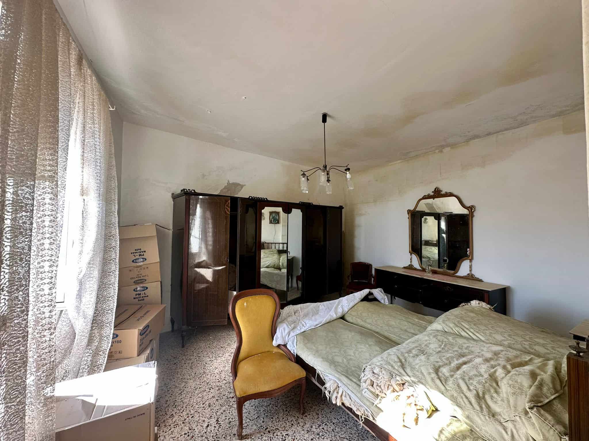 Vecchio soggiorno con mobili vintage, letto sfatto, grande specchio antico, armadio in legno scuro, poltrona gialla in stile classico, pareti leggermente ammuffite, ambiente luminoso con tende in pizzo.