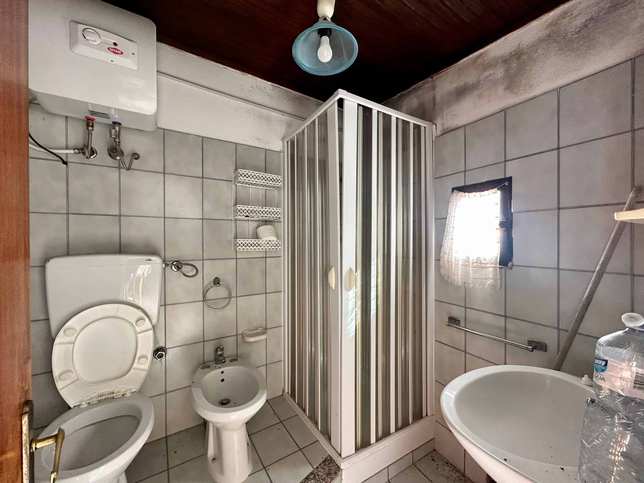 Doccia in bagno con pareti in piastrelle, toilette e bidet, finestra con tende, lavandino, acqua calda e fredda, ambiente semplice e funzionale, situato in un'abitazione italiana.