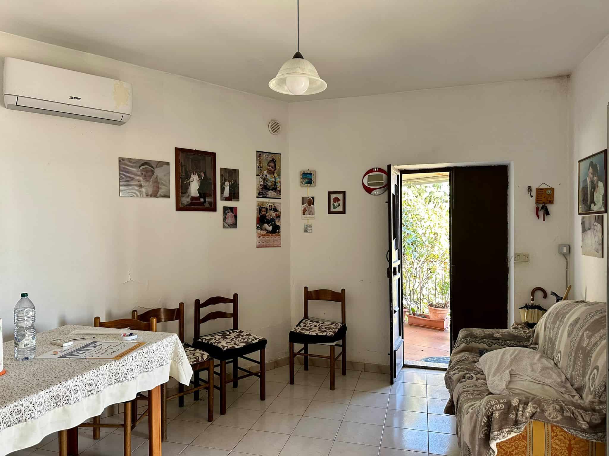 Soggiorno accogliente con parete decorata da foto di famiglia, tavolo con tovaglia ricamata, sedie in legno e divano con copertura elegante, ingresso con porta aperta su terrazza con piante verdi, aria condizionata e luce naturale.