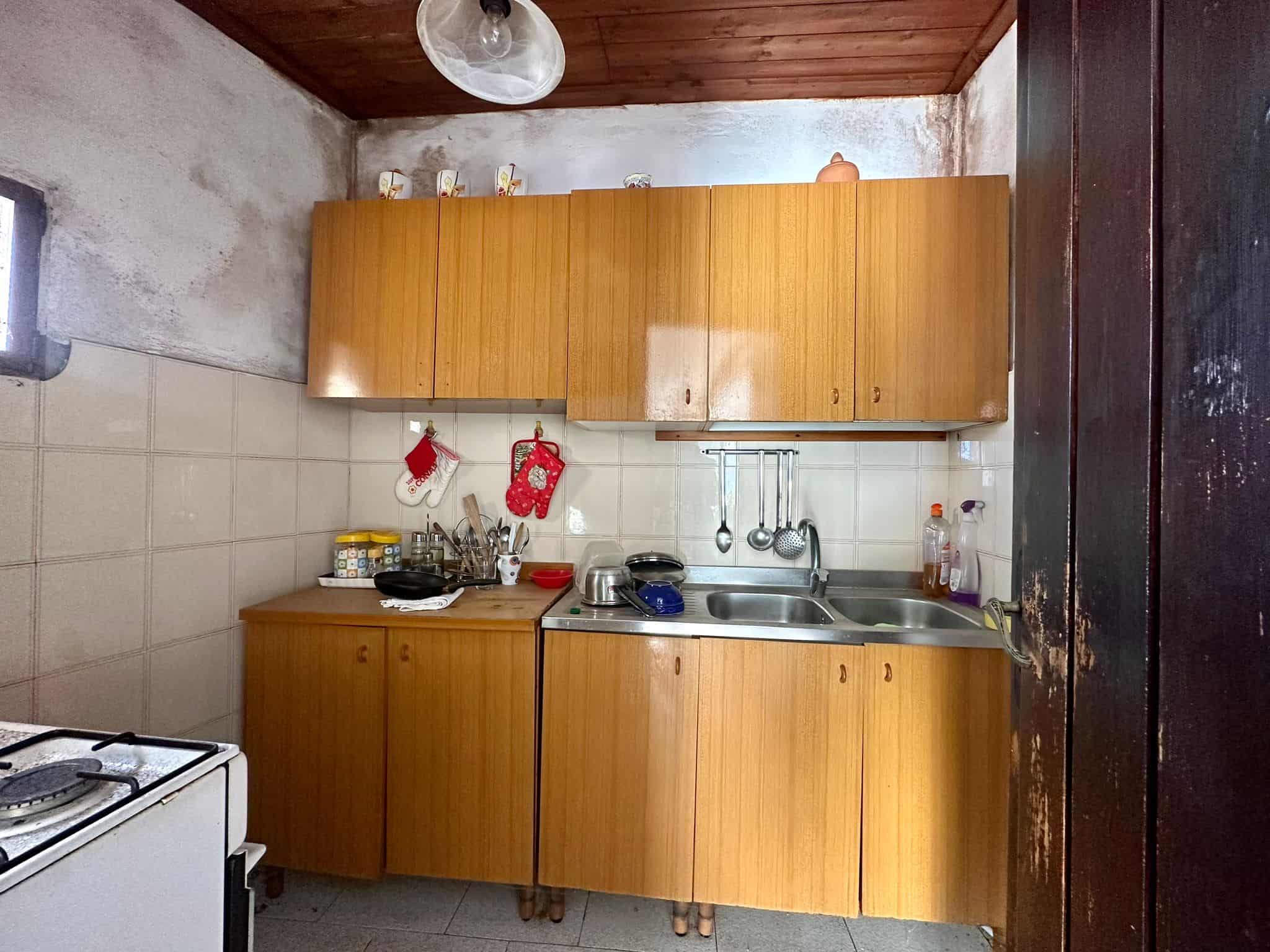 Cucina cucinino rustico in stile anni 70 con mobili in legno chiaro, lavabo doppio e dettagli nostalgici, ideale per case di campagna o appartamenti tradizionali italiani.