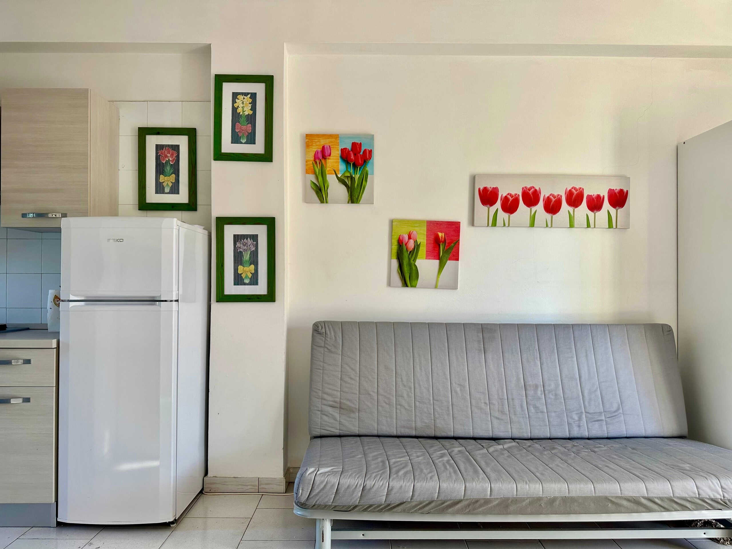 Fondo soggiorno con divano letto grigio, parete decorata con quadri di tulipani rossi e altri fiori in cornici verdi, accanto a frigorifero bianco e cucina ad angolo, ambiente accogliente e luminoso.