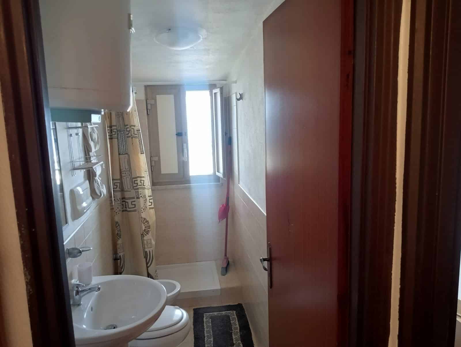 Bagno di appartamento con finestra, lavabo con doppio lavandino, WC, asciugamani, pavimento in piastrelle chiare, spazzola per pavimenti e tende decorative. Spazi funzionali e luminosi.