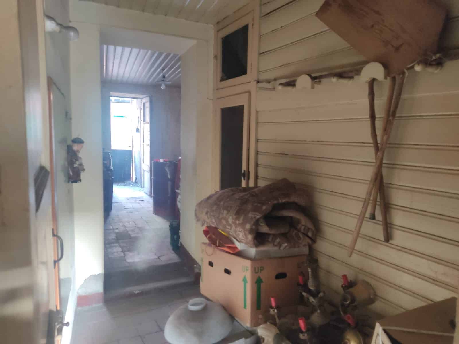 Finestra in una stanza con pareti rivestite di pannelli di legno e oggetti come pullover, box con arnesi e bottiglie di vino. Ambiente rustico e funzionale, ideale per uso casalingo o magazzino.