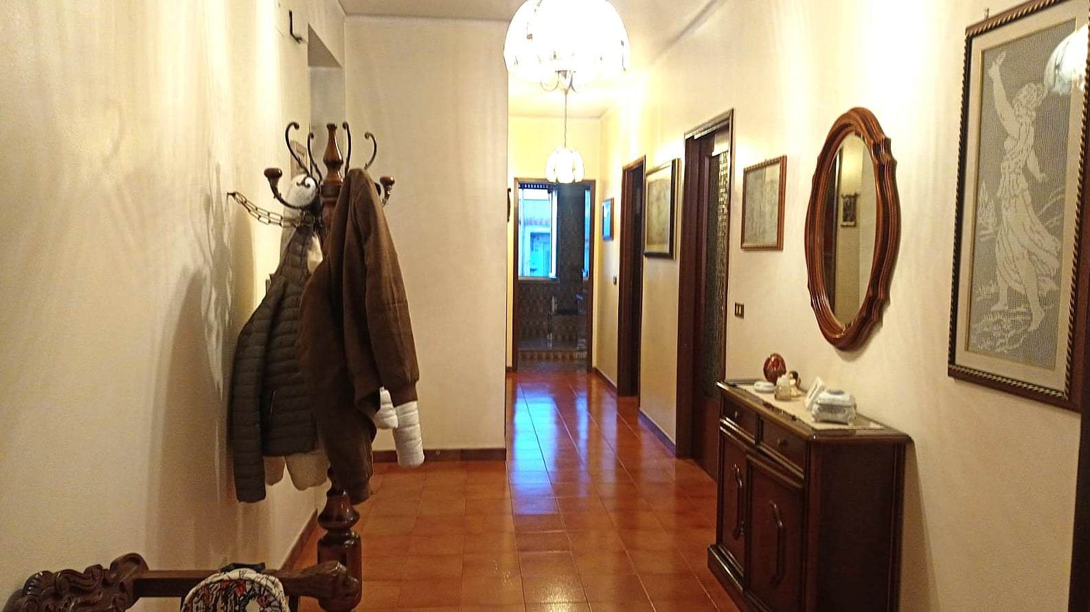 Vestibolo arredato con arredi classici, specchio in legno e pareti decorate, corredo di cappotti, quadri alle pareti e illuminazione calda, ingresso elegante di una casa NextCasa.