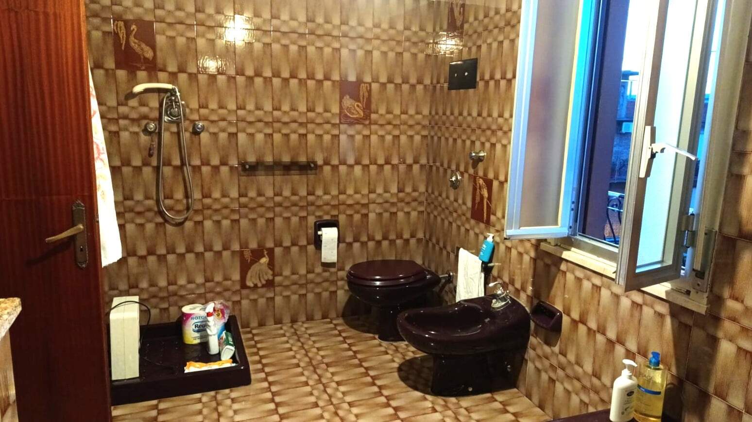 Piastrelle bagno stile vintage con piastrelle marroni e decori artistici, wc e bidet scuri, finestra aperta per aerazione, setup funzionale e decorativo, ideale per bagno classico o retrò.