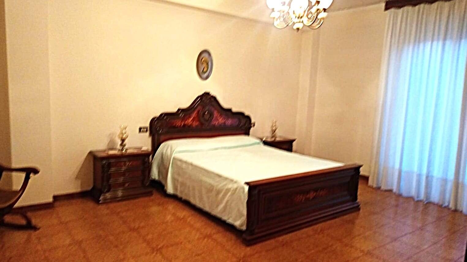 Letto matrimoniale in una camera elegante con arredi classici, illuminate da una lampada a soffitto, con tende bianche e pareti neutre, ideale per soggiorno confortevole e di lusso.