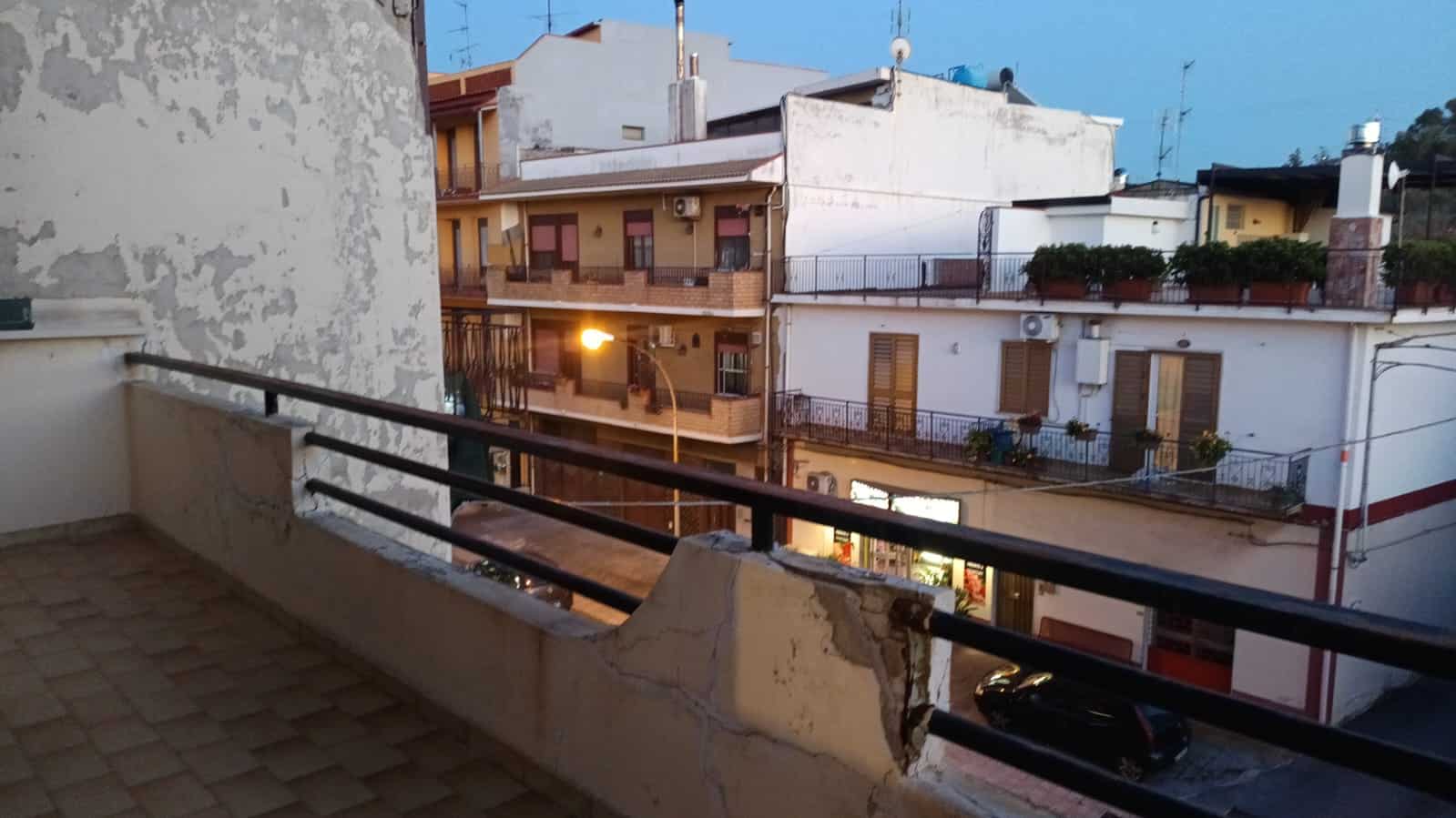 Balcone di un appartamento in zona urbana con vista sulla strada e altri edifici residenziali, in stile città, al tramonto, con luci accese e piante sui balconi.