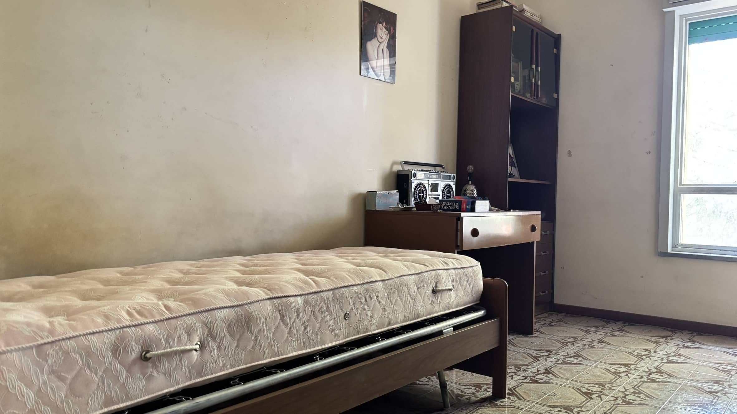 Letto singolo con materasso vecchio in una stanza semplice e spoglia, con mobile bagno e scaffale in legno, finestra luminosa e pavimento in piastrelle geometriche.