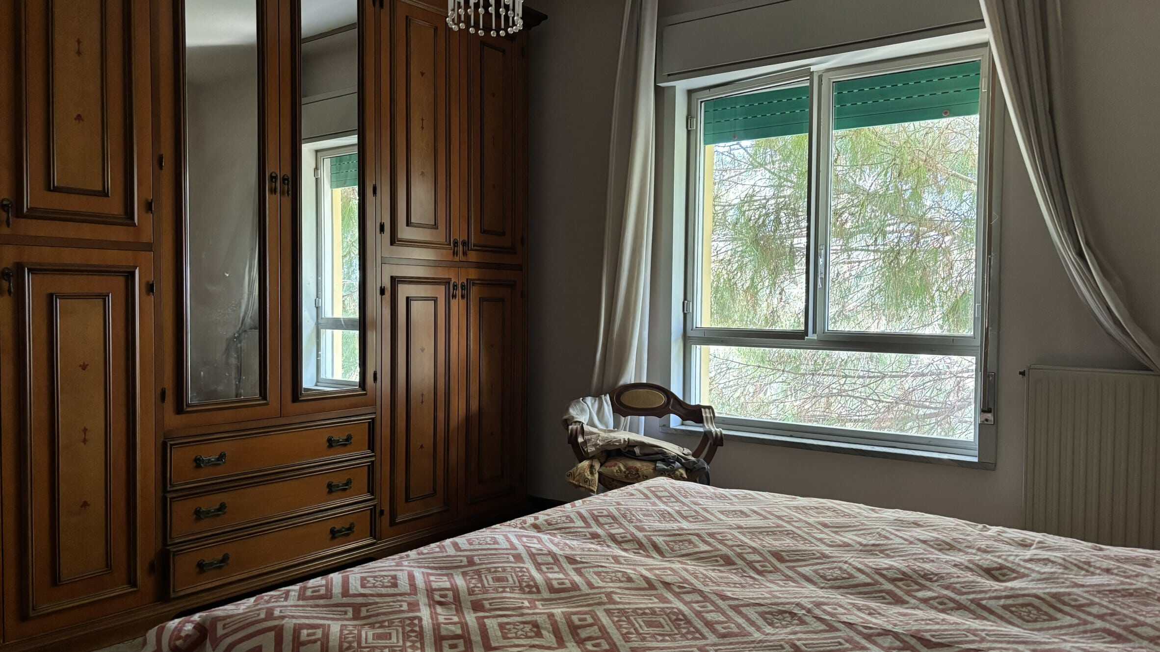 Armadio in legno massello con ante a battente e cassetti, vista da una camera da letto luminosa con grandi finestre e tende bianche. Esempio di arredo classico e funzionale per case e appartamenti.