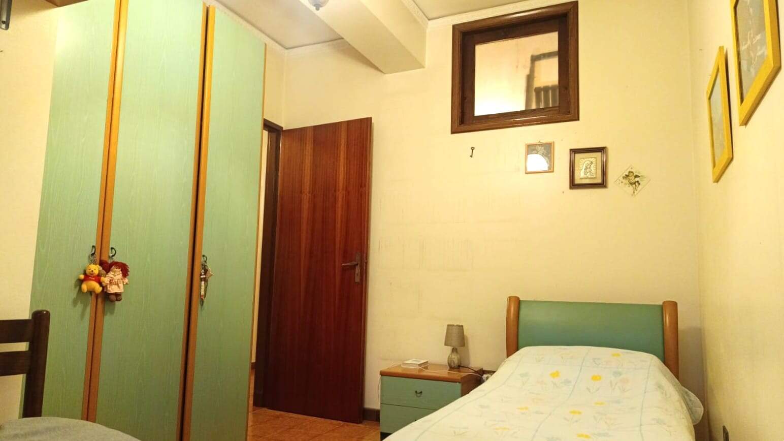Armadio a muro verde pastello con ganci decorativi, camera da letto accogliente con pareti gialle, letto singolo con copriletto di fantasia, comò con lampada e quadri alle pareti, finestra con cornice di legno.
