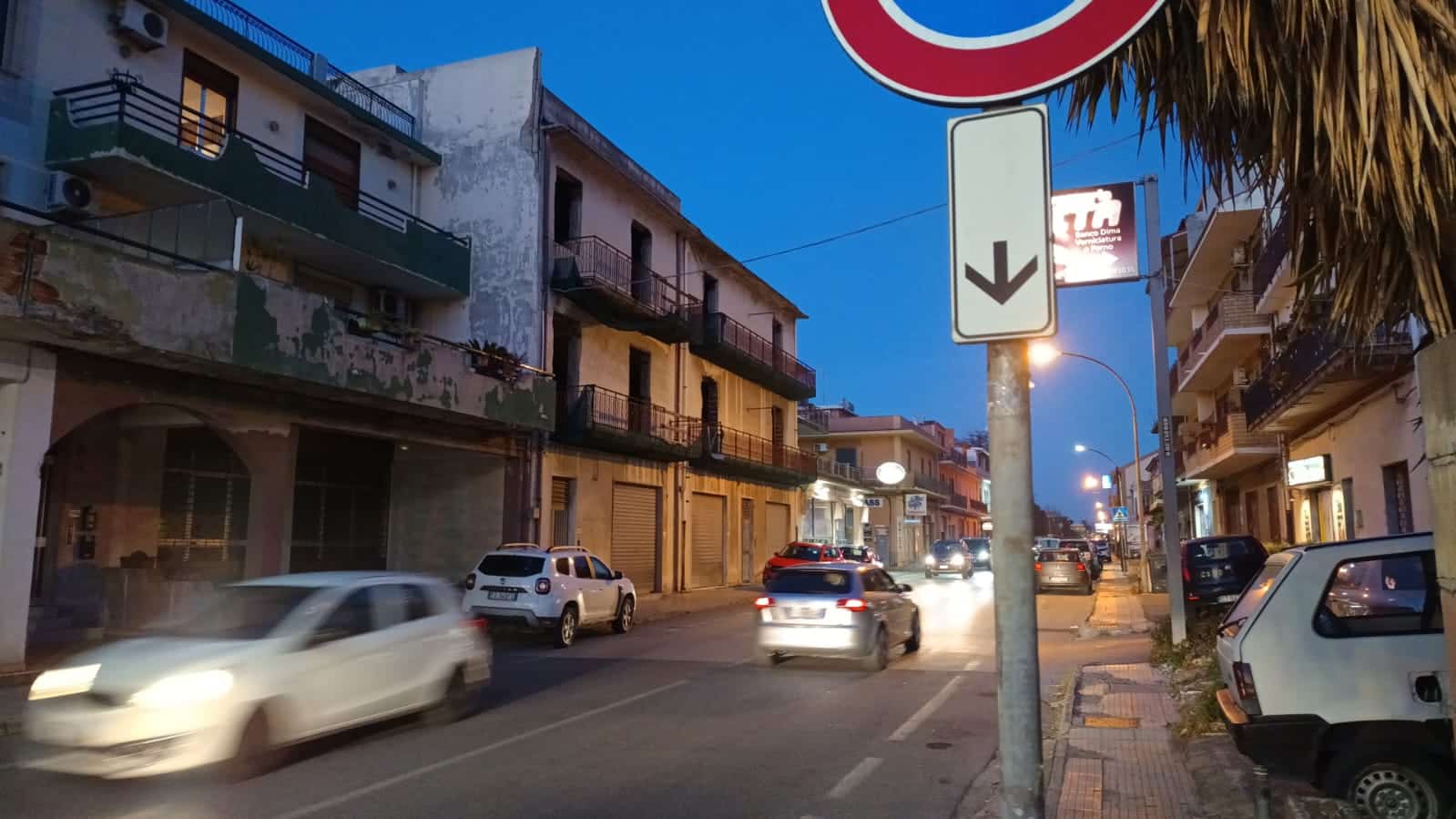 Auto in movimento sulla strada di notte, con edifici residenziali e negozi, segnale stradale e illuminate pubbliche, scena urbana per immobili e investimenti immobiliari NextCasa.