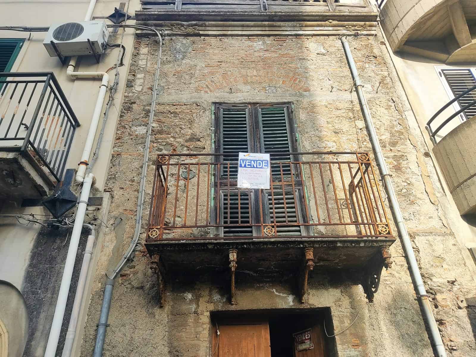 Balcone antico su edificio storico con "Vende" cartello immobiliare, caratteristiche di un'unità residenziale da ristrutturare, facciata in pietra e ferro battuto.