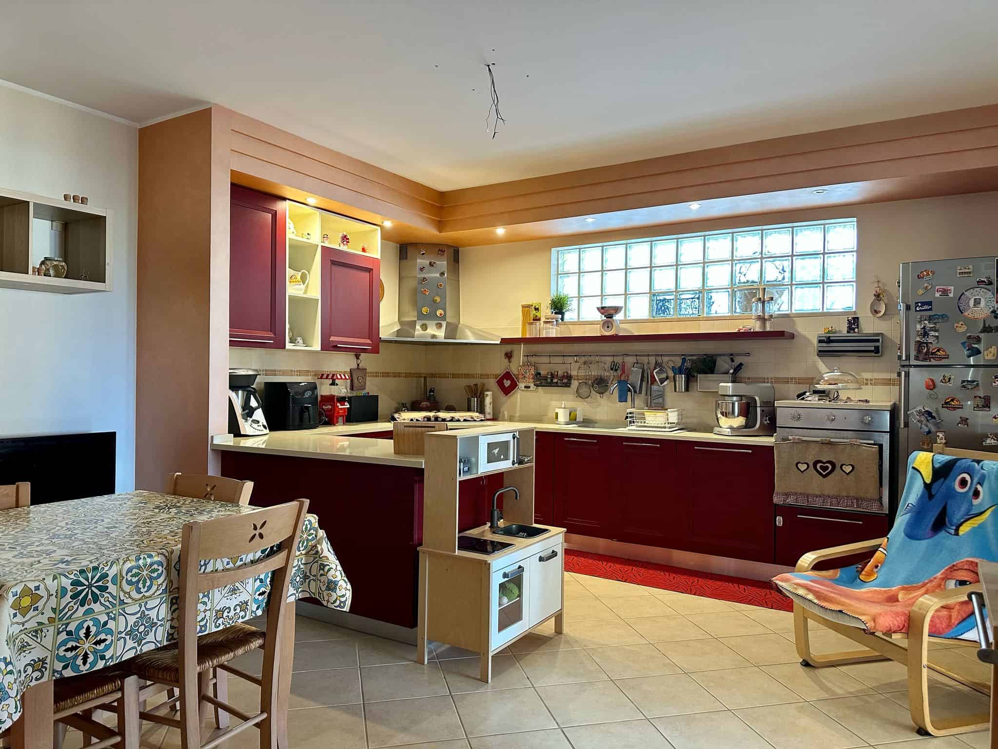 Luminoso cucina moderna con mobili color vinaccia, piano cottura a gas, frigorifero decorato con magneti, e spaziosa zona pranzo con tavolo e sedie in legno, ideale per ambienti abitativi dinamici e accoglienti.