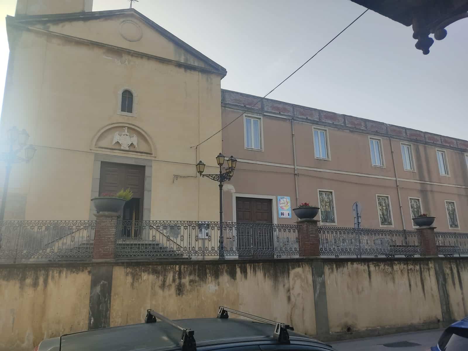 Vecchia chiesa con facciata semplice e dettaglio di rilievo sulla porta, in un contesto urbano. Edificio storico con elementi architettonici tradizionali italiani, situato in una zona con pavimentazione e arredi pubblici.