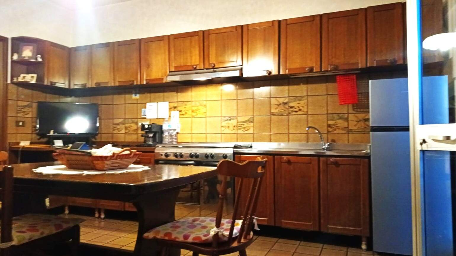 Fornello a gas in cucina con ante in legno, pareti rivestite di piastrelle color marrone, tavolo in legno con sedie con imbottitura decorata, frigorifero blu e schermo TV, ambiente rustico e caldo.