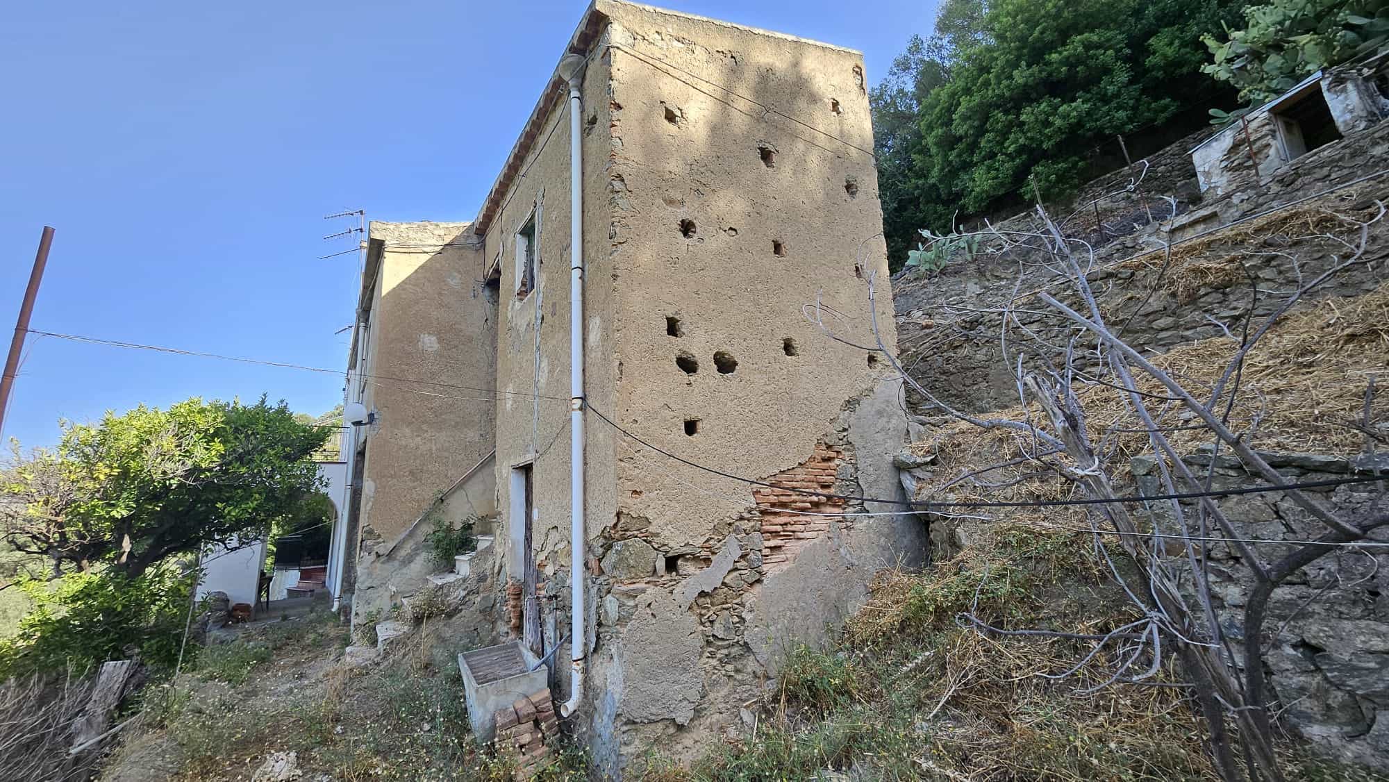 Vecchio edificio in rovina con pareti danneggiate e fori, situato su un pendio collinare con vegetazione, pronto per ristrutturazione, in un contesto di natura e storia.