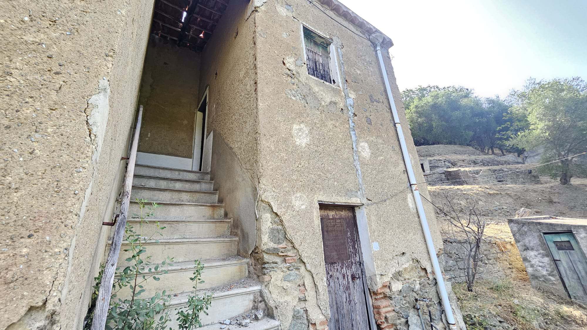 Vecchio edificio in pietra con scala esterna e portone di legno, situato in zona rurale con vegetazione e alberi circostanti, perfetto per ristrutturazione e investimenti immobiliari.