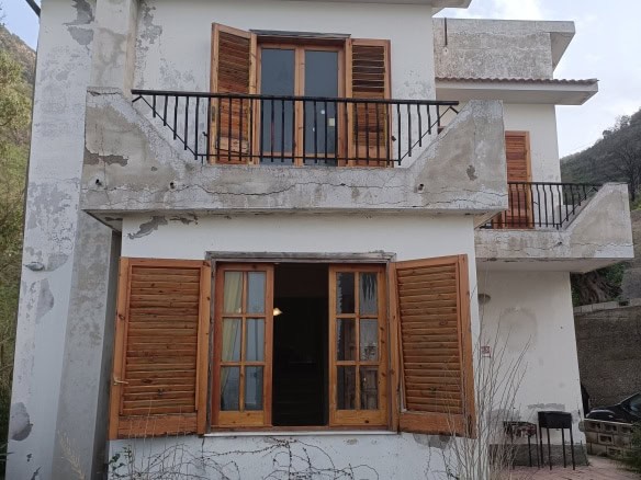Edificio residenziale con facciata in condizioni di ristrutturazione parziale, con finestre in legno e balconi in ferro battuto, ambienti esterni con giardino e vista montagna, stile autentico e rustico.