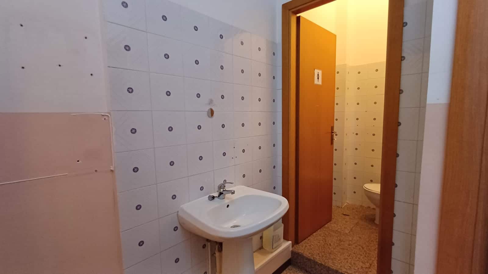 Lavabo in bagno con pareti rivestite di piastrelle bianche a motivo floreale, porta in legno che dà accesso a un wc separato, ambiente funzionale e ottimale per la ristrutturazione immobiliare.
