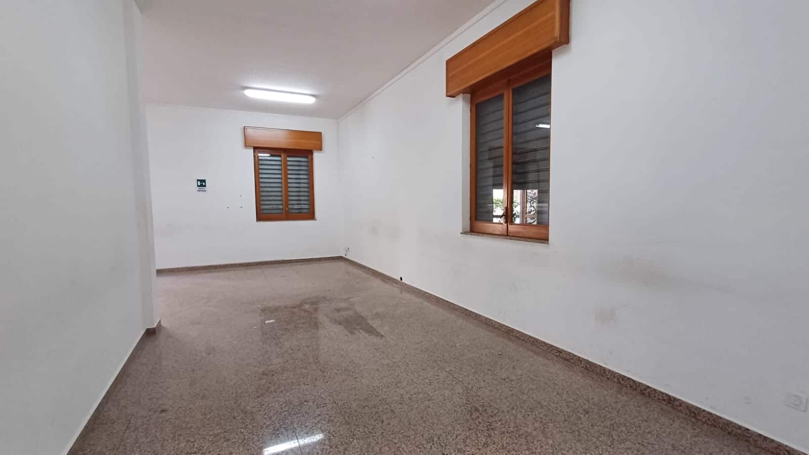 Spazio interno vuoto di appartamento con pareti bianche, pavimento in gres e finestre in legno con persiane, ideale per ristrutturazione o affitto.