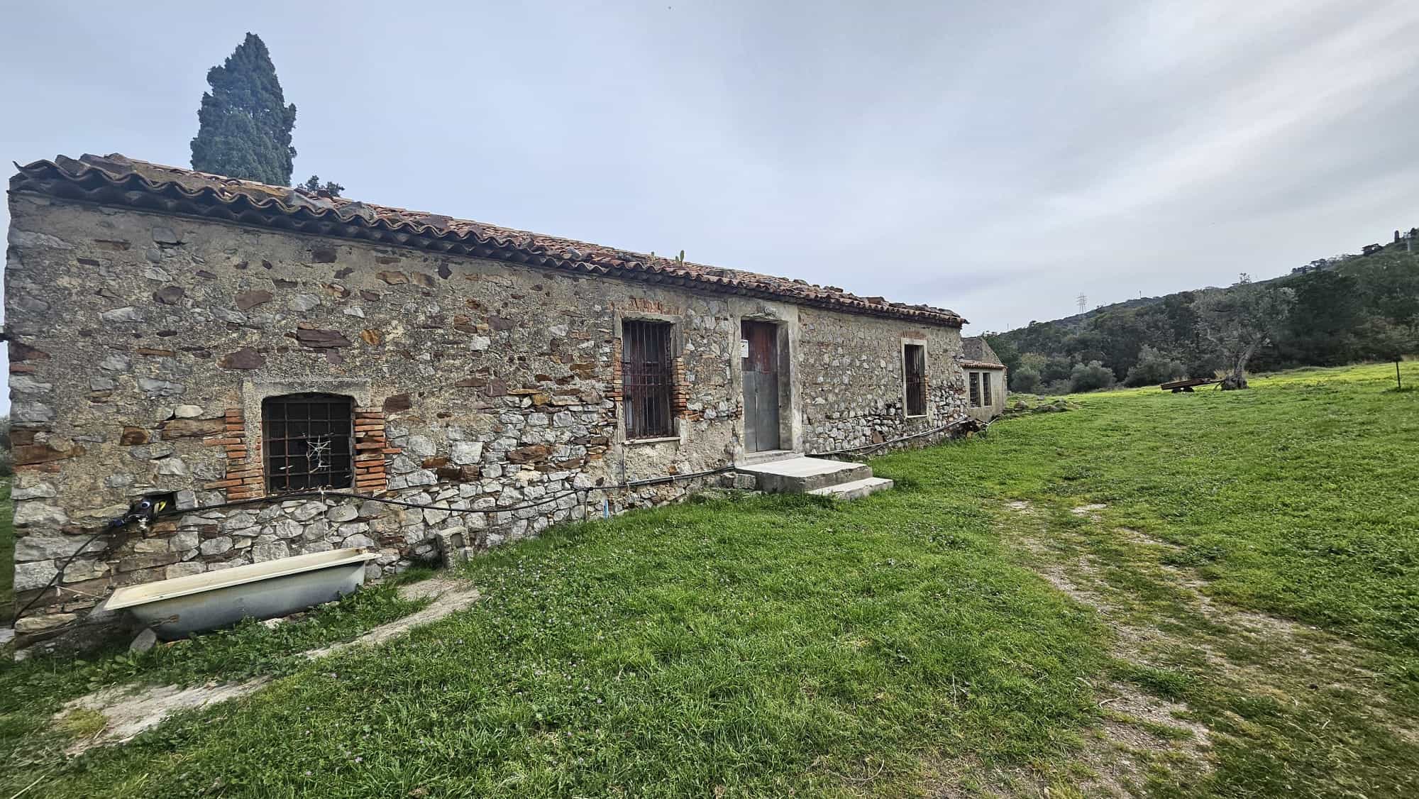 Vecchio casolare in pietra con cancello in metallo, immerso nel verde della campagna toscana, ideale per ristrutturazione e sviluppo immobiliare, con vista sulle colline e ambiente rurale.