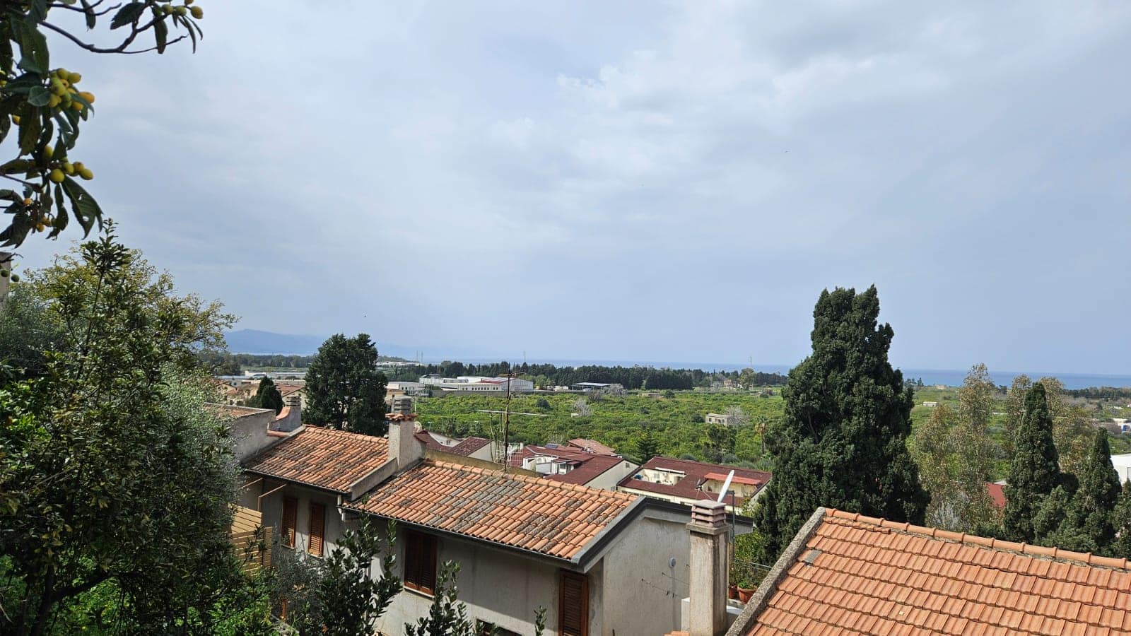 Balcone con vista panoramica su case con tetti in terracotta e paesaggi verdi, vista mare e colline in lontananza, cielo nuvoloso, atmosfera tranquilla e mediterranea.