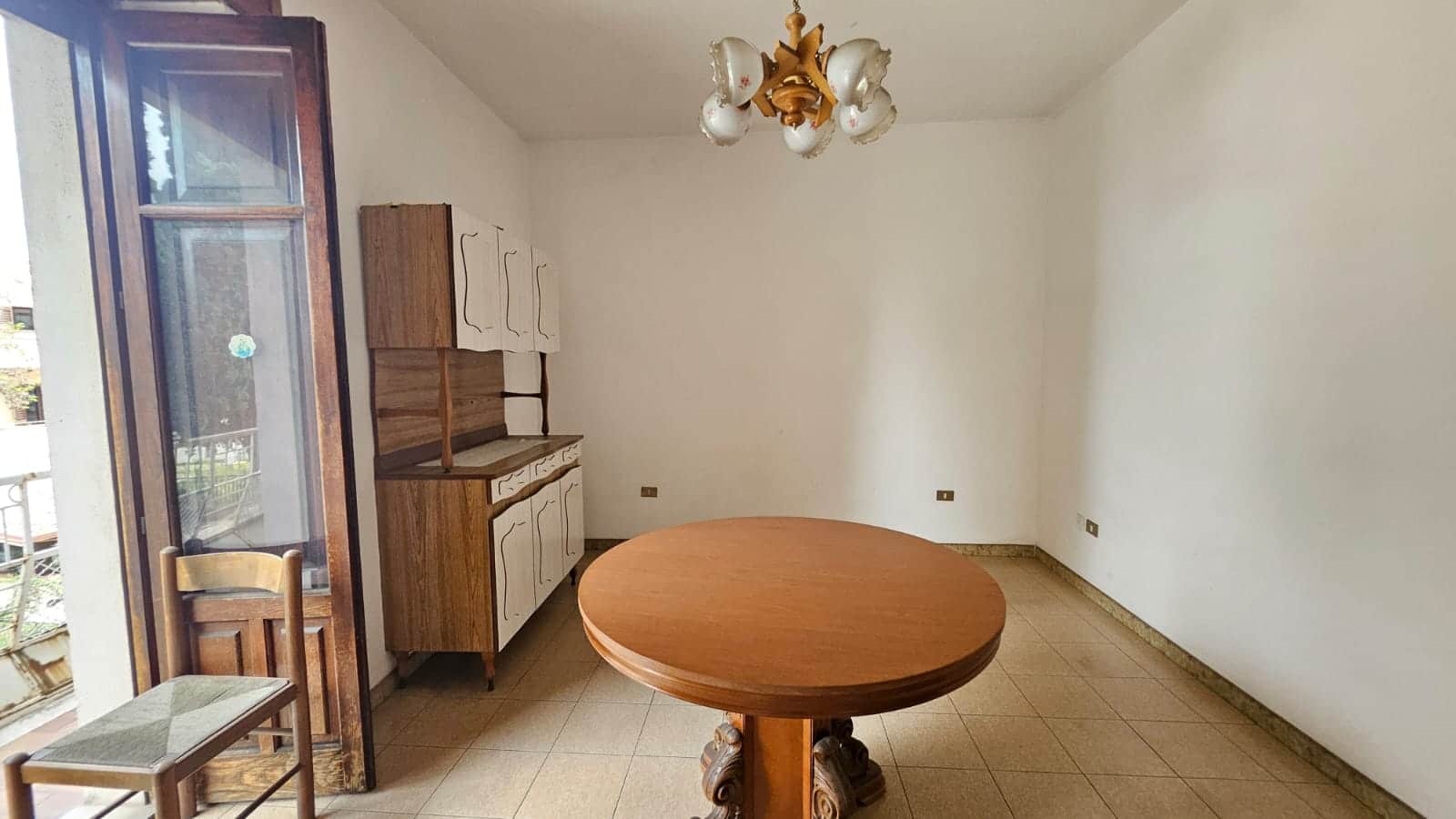 Soggiorno con pareti bianche, tavolo rotondo in legno, mobile in stile vintage e cucina a vista, illuminato da lampadario d'epoca, balcone con accesso in legno e sedie in stile classico.