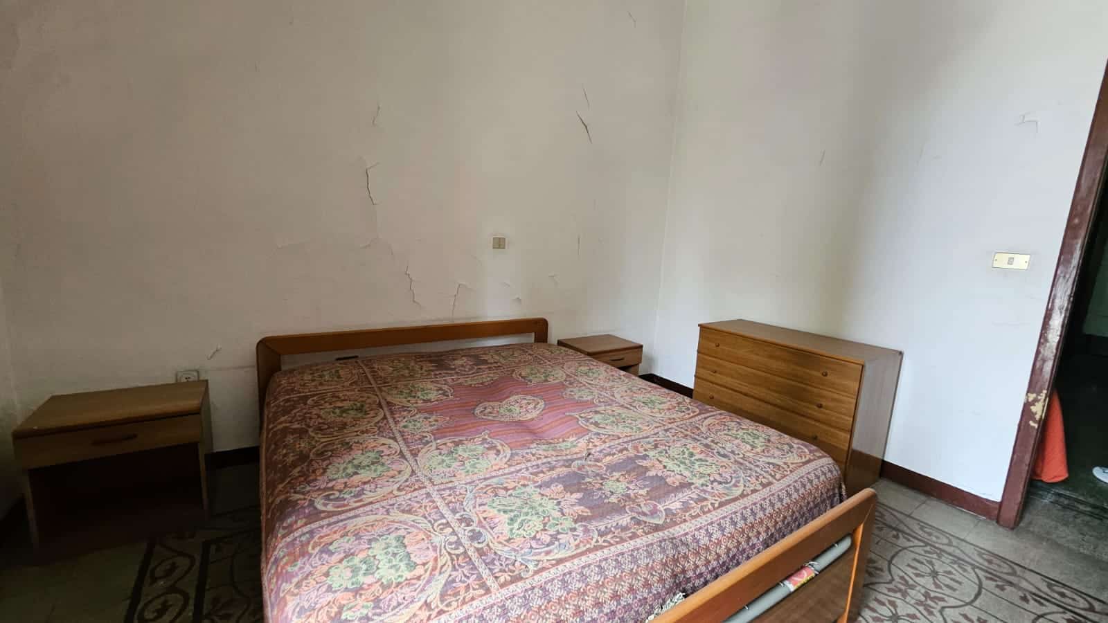 Letto matrimoniale con testiera in legno, letto singolo e comodini in camera da letto. Pareti con fessure e crepe visibili, pavimento in piastrelle con motivo decorativo, stanza semplice e funzionale.