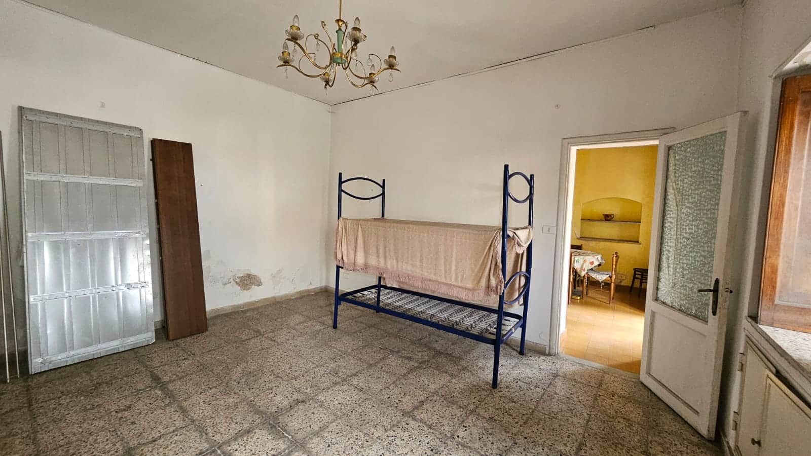 Vecchia stanza abitabile con soffitto decorato, armadietto metallico, letto a castello senza materasso, pareti bianche con segni di umidità, arredamento semplice, ideale per ristrutturazione o investimento immobiliario.
