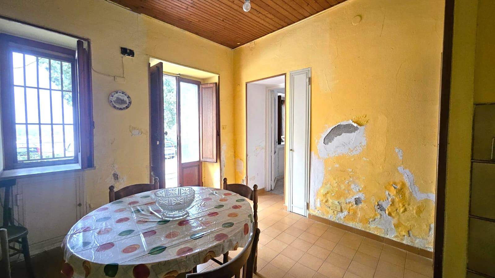 Vecchia cucina con pareti gialle e soffitto in legno, tavolo rotondo con tovaglia colorata, finestra con grate e ingresso alla balconata, dettagli di ristrutturazione e impianto elettrico a vista.