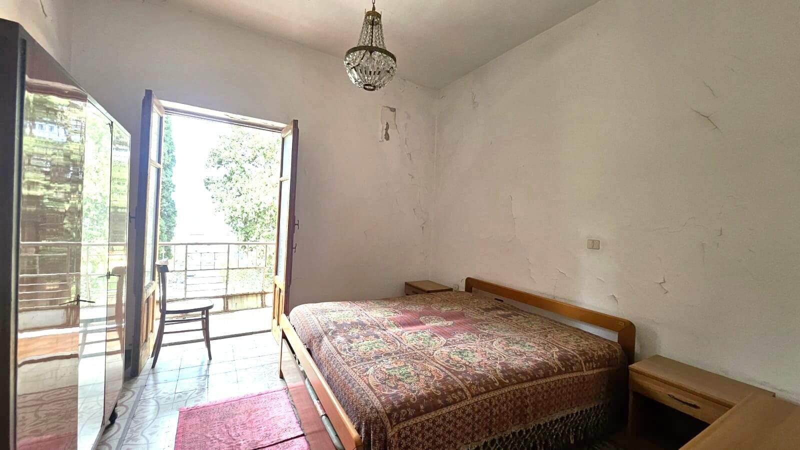 Vecchia camera da letto con pareti screpolate e vetri aperti che portano a un balcone, arredamento semplice, con letto in legno, tappeto rosso, lampadario di cristallo, atmosfera di casa da rinnovare.