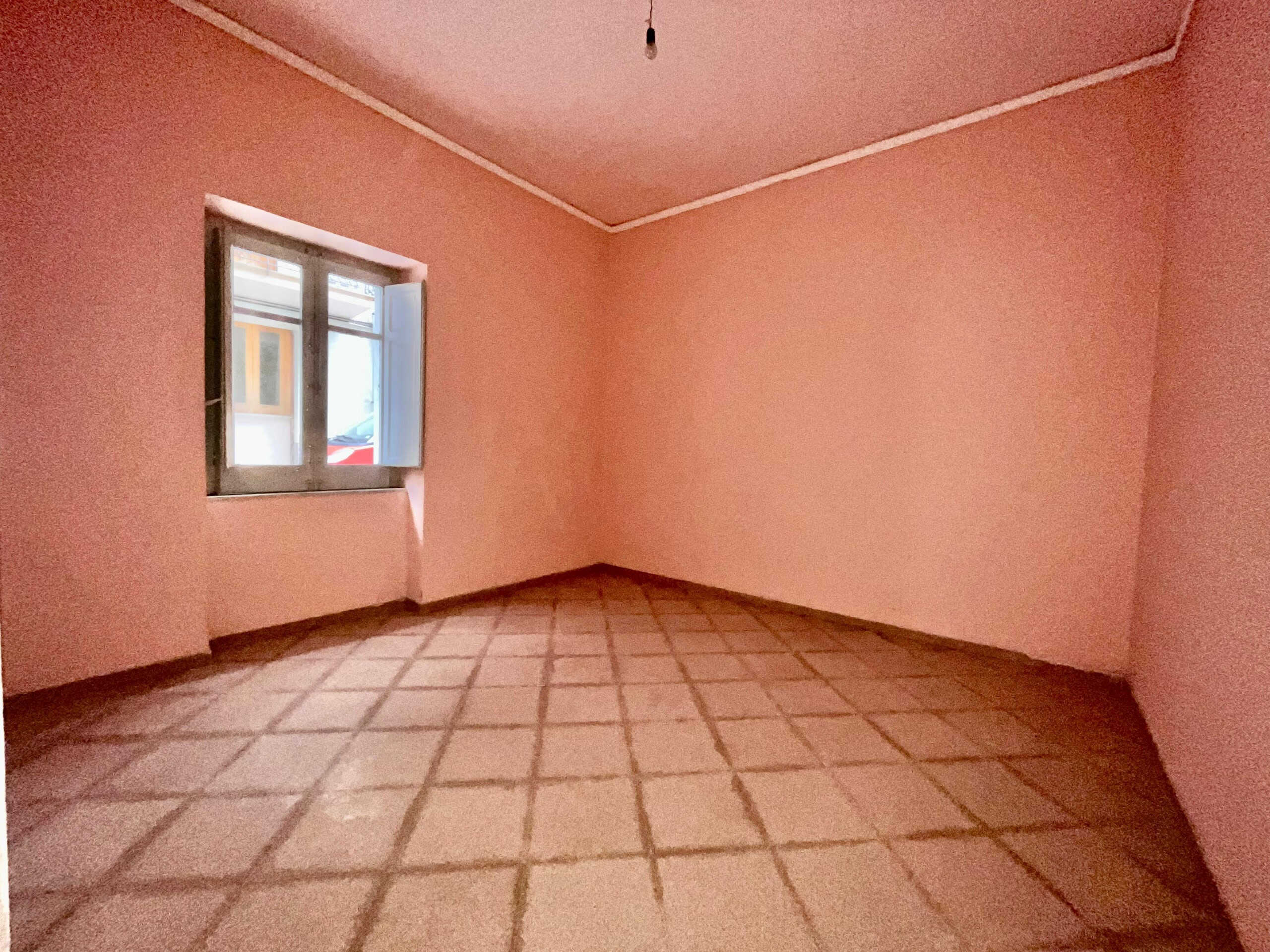 Stanza vuota con pareti rosa e pavimento in piastrelle, finestra con persiana bianca, ambiente luminoso e spazioso, perfetto per ristrutturare e personalizzare per la vendita o l'affitto.