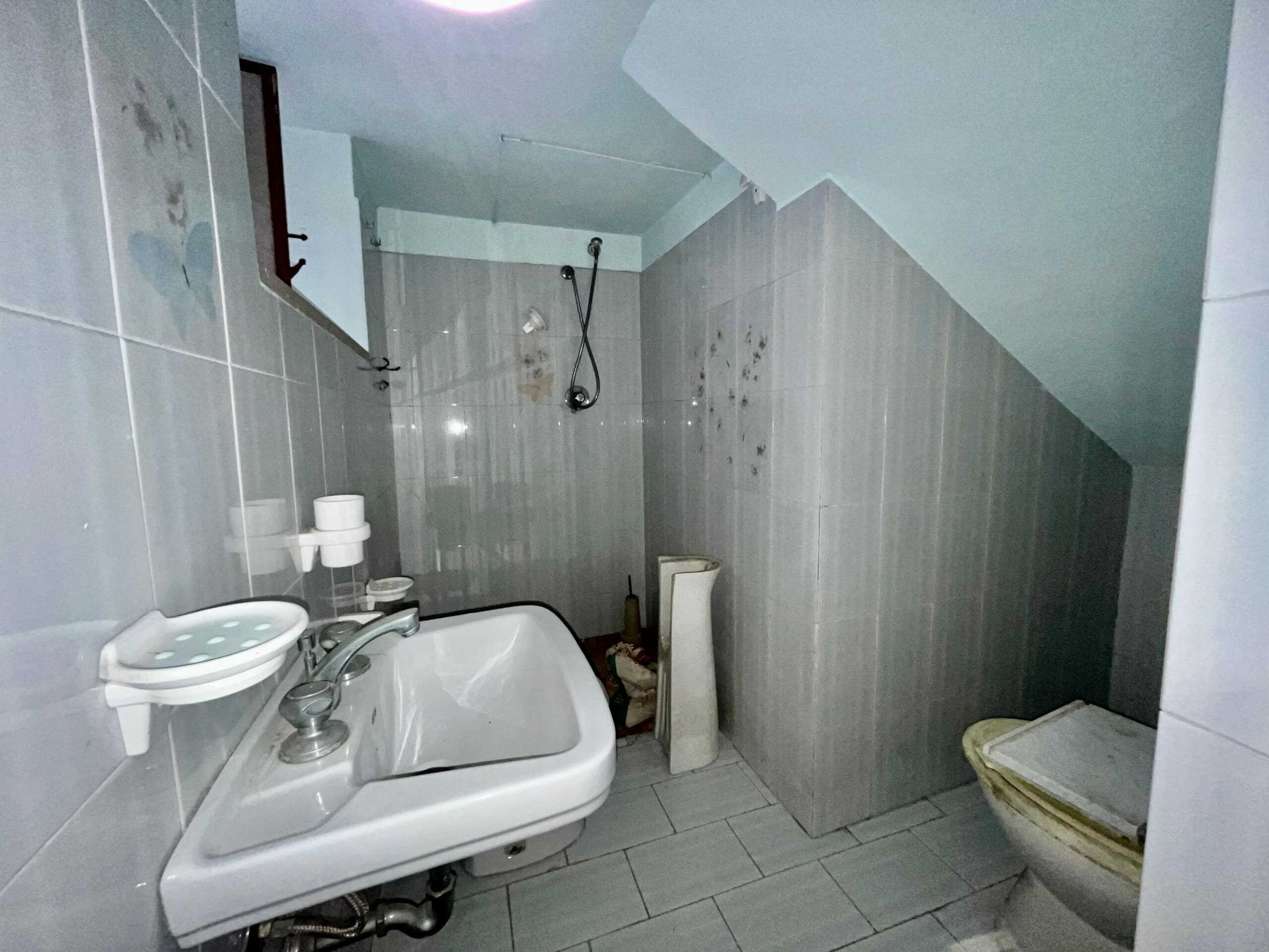 Lavabo e servizi igienici in un bagno in condizioni precarie con perdite e muffa, arredato con accessori di base, pareti rivestite in piastrelle grigie e pavimento in piastrelle coordinate.
