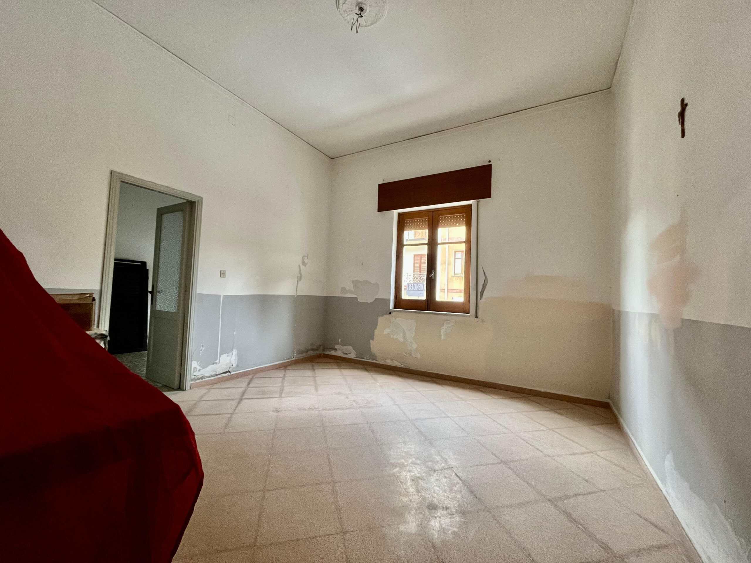 Vista di una stanza vuota in fase di ristrutturazione, pareti con vernice parziale e pavimento in piastrelle. Finestra che lascia entrare luce naturale, soffitto con lampadario, ambiente ideale per nuove personalizzazioni edilizie.