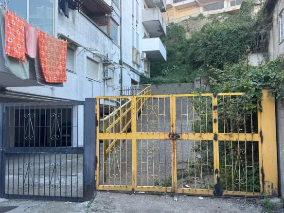 Cancelletto in metallo giallo con passaggio pedonale, ingresso di un edificio residenziale in zona urbana, con vegetazione e pareti di palazzi circostanti.