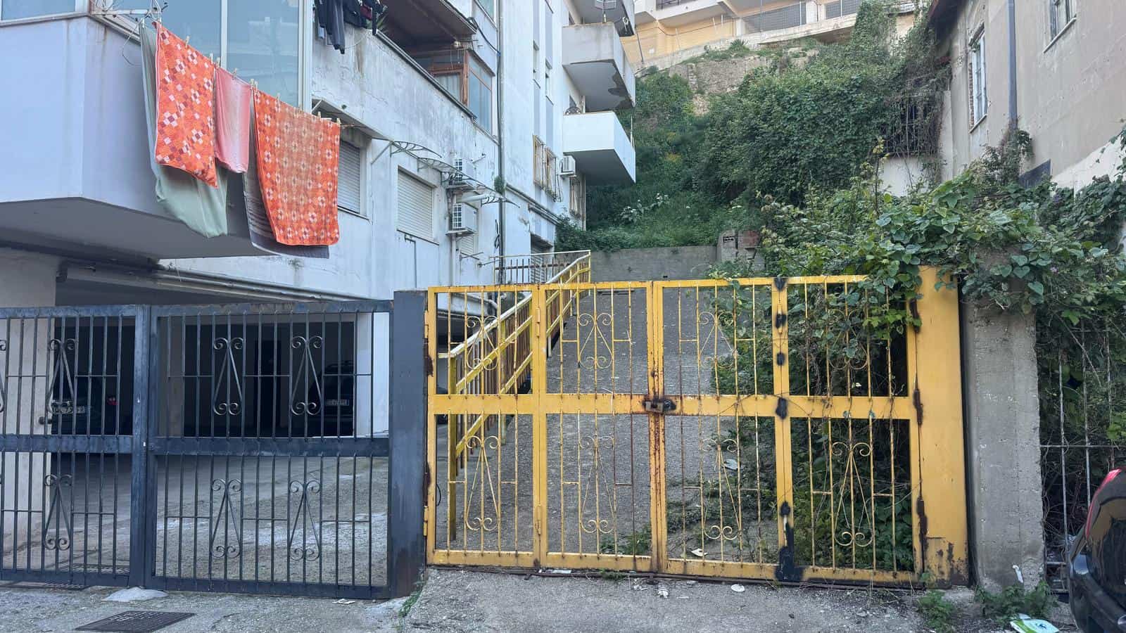a4007459-f6bd-457a-96cc-d1dd3bf17d57 Cancelletto in metallo giallo con passaggio pedonale, ingresso di un edificio residenziale in zona urbana, con vegetazione e pareti di palazzi circostanti.