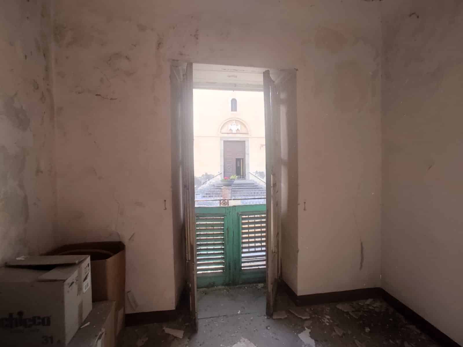 Finestra con vista in una stanza in corso di ristrutturazione, pareti grezze e pavimento sconnesso, ambiente luminoso e spazioso con prospettiva sulla chiesa storica all'esterno, pronto per essere arredato.