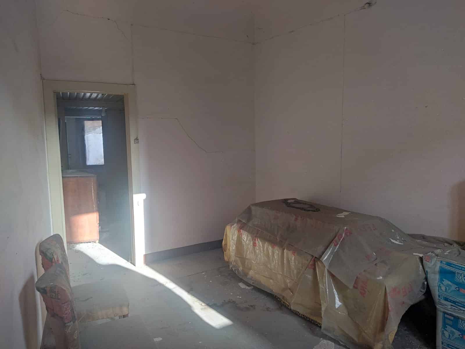 S fronte di una stanza in ristrutturazione, pareti e pavimenti da rifare, coperto con teli protettivi, porta aperta che rivela uno spazio in fase di lavori, ambiente luminoso e in attesa di personalizzazione.